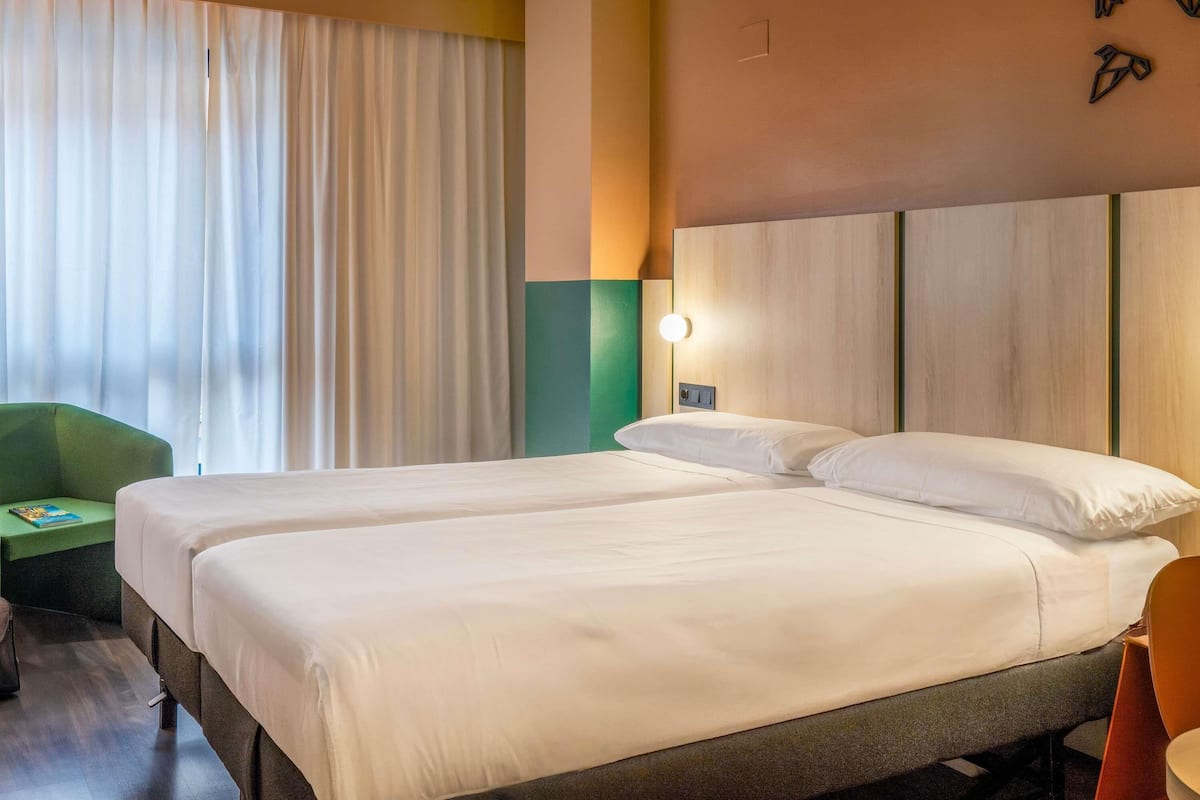 Hotellihuone, jossa on kaksi sänkyä, vihreä tuoli ja seinään kiinnitetty valaisin.