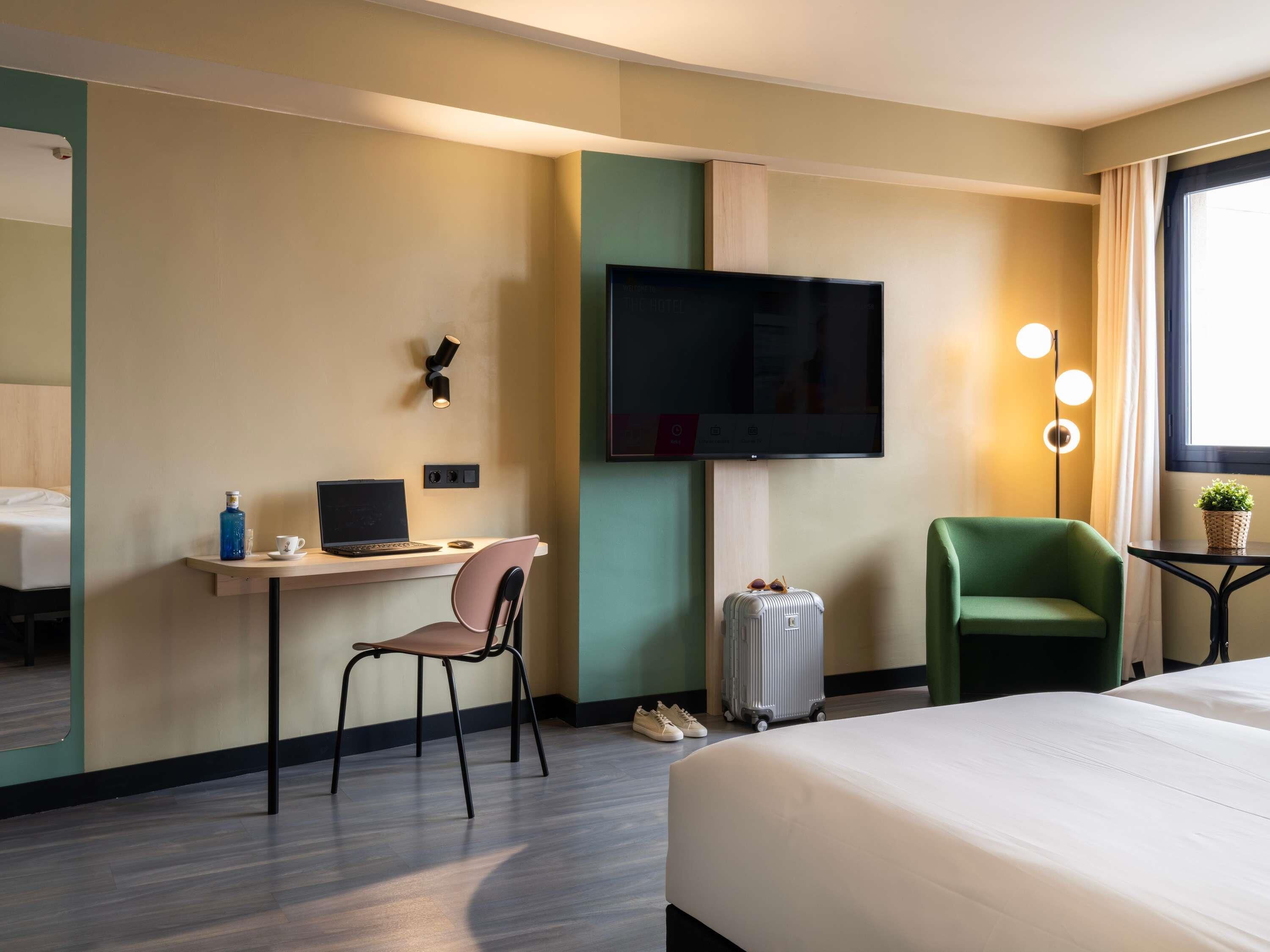 A hotel room with a bed, a desk with a laptop, a chair, a suitcase, and a television.