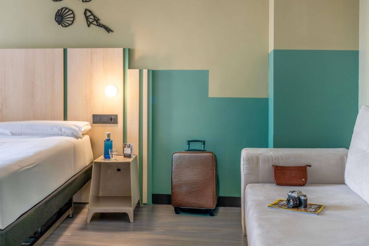 A hotel room with a bed, a nightstand, a suitcase, a sofa, and a wall with decorative elements.