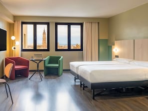 A hotel room with a bed, two chairs, a desk with a laptop, and large windows with a city view.