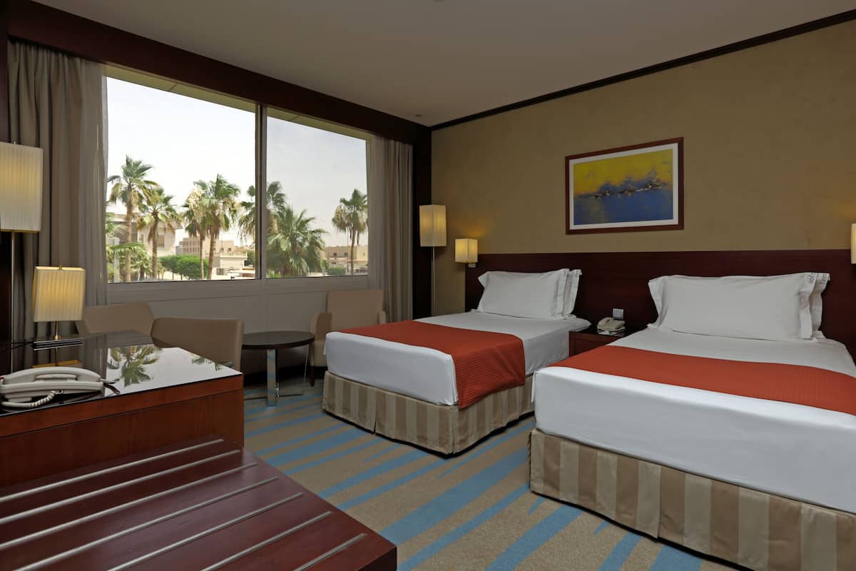 A hotel room with two beds, a desk, a chair, and a window with a view of palm trees.