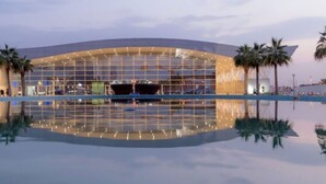 A modern hotel with a large glass facade, palm trees, and a pool reflecting the building.