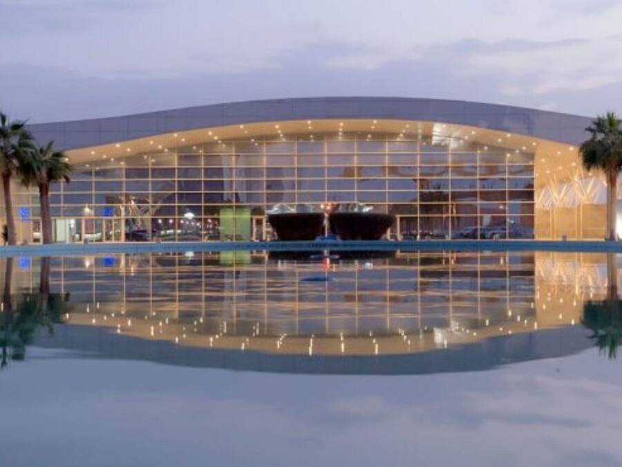 Un hotel moderno con una amplia fachada de vidrio, palmeras y una piscina que refleja el edificio.