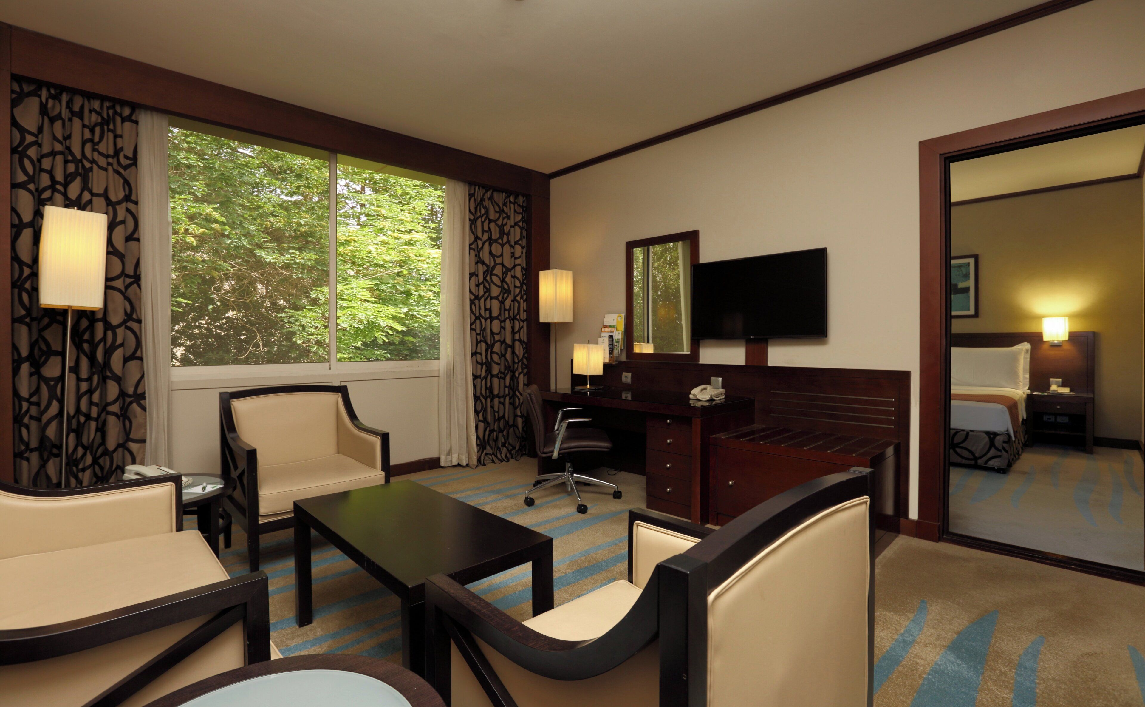 A hotel room with a desk, two chairs, a television, and a large window with a view of trees.