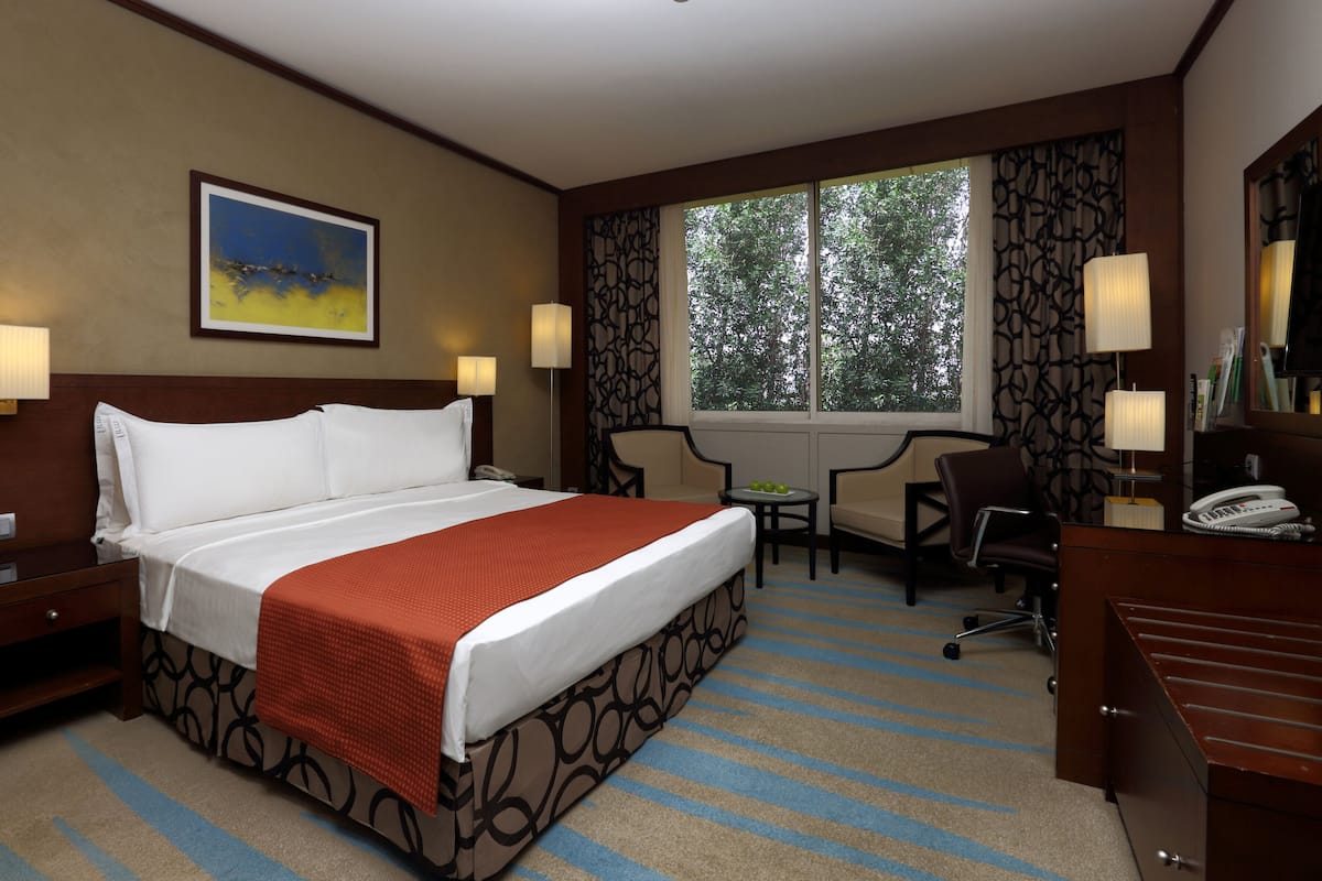 A hotel room with a large bed, a desk, two chairs, a window with a view of trees, and a painting on the wall.