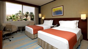 A hotel room with two beds, a desk, a chair, and a window with a view of palm trees and buildings.