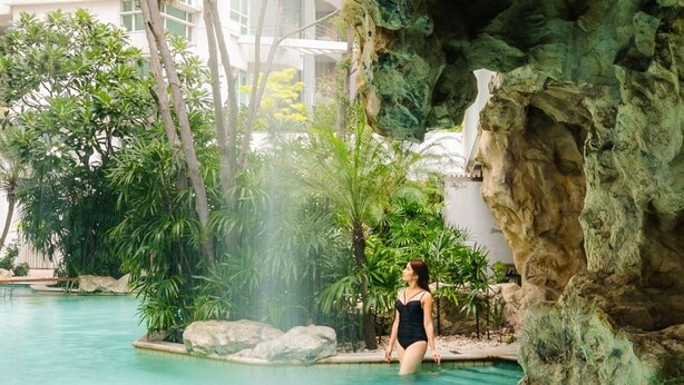 A woman in a black swimsuit standing by a pool with a rocky cave backdrop.