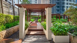 A covered walkway with a wooden bench, surrounded by greenery and a stone wall.