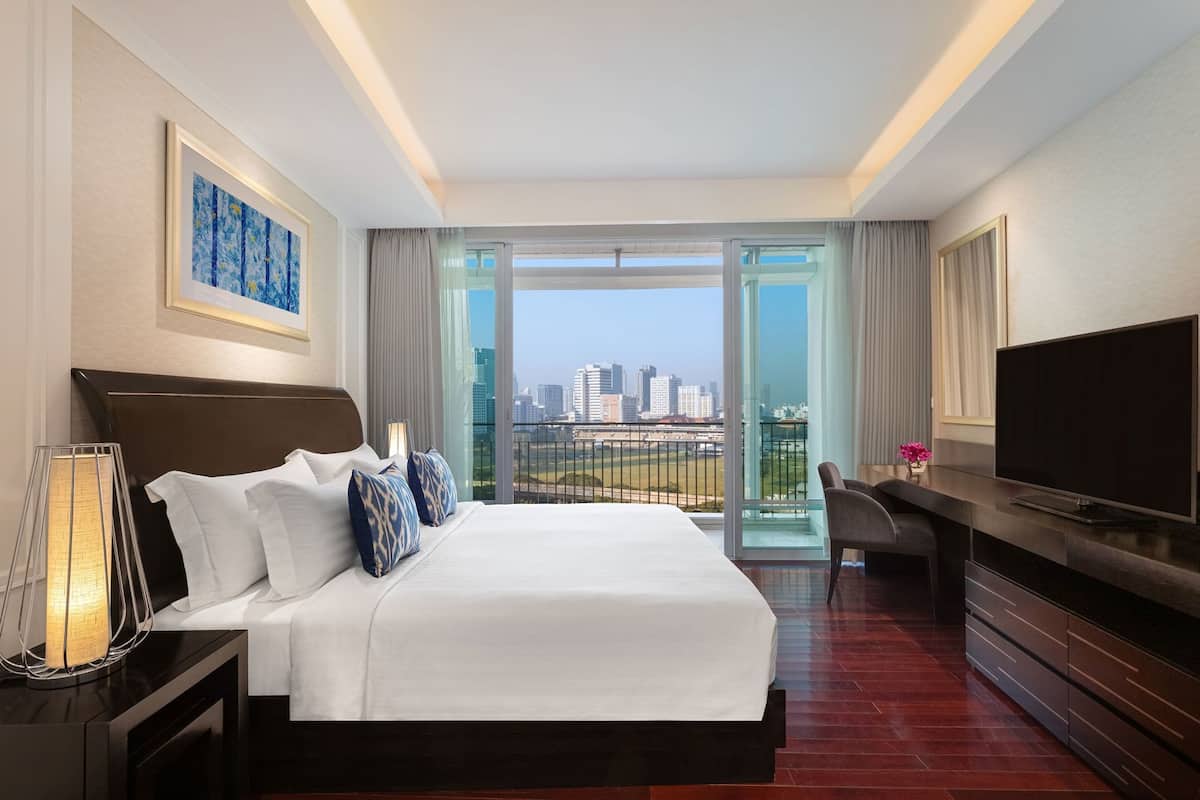 A hotel room with a large bed, a desk with a TV, a view of the city, and a balcony.