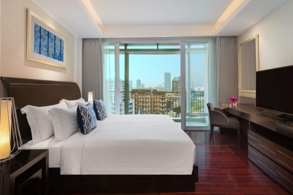 A hotel room with a large bed, a desk, a television, and a view of the cityscape through large windows.