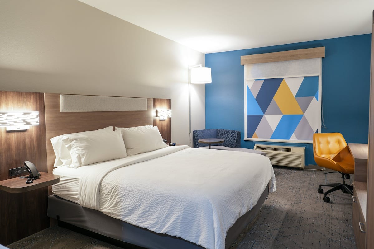 A hotel room with a large bed, bedside table with a phone, a chair, a desk, and a wall with geometric artwork.
