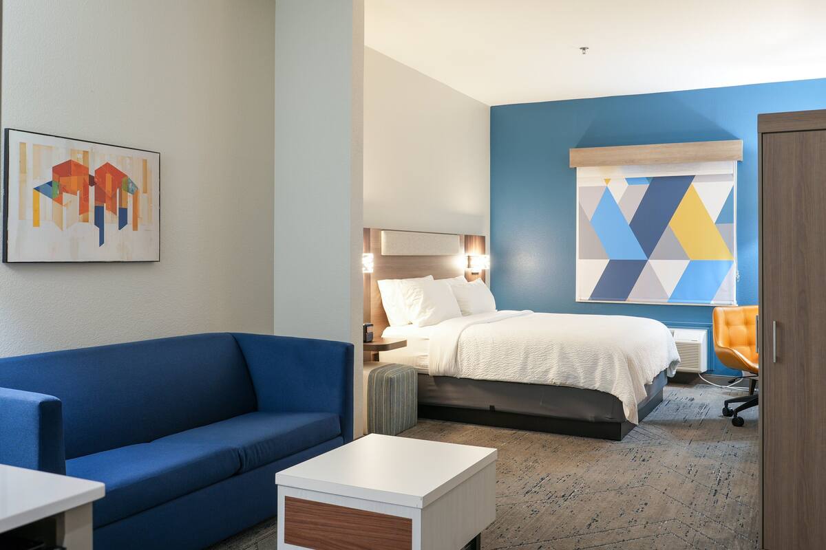 A modern hotel room with a blue wall, a bed with white bedding, a blue sofa, a small white table, and a colorful abstract painting.
