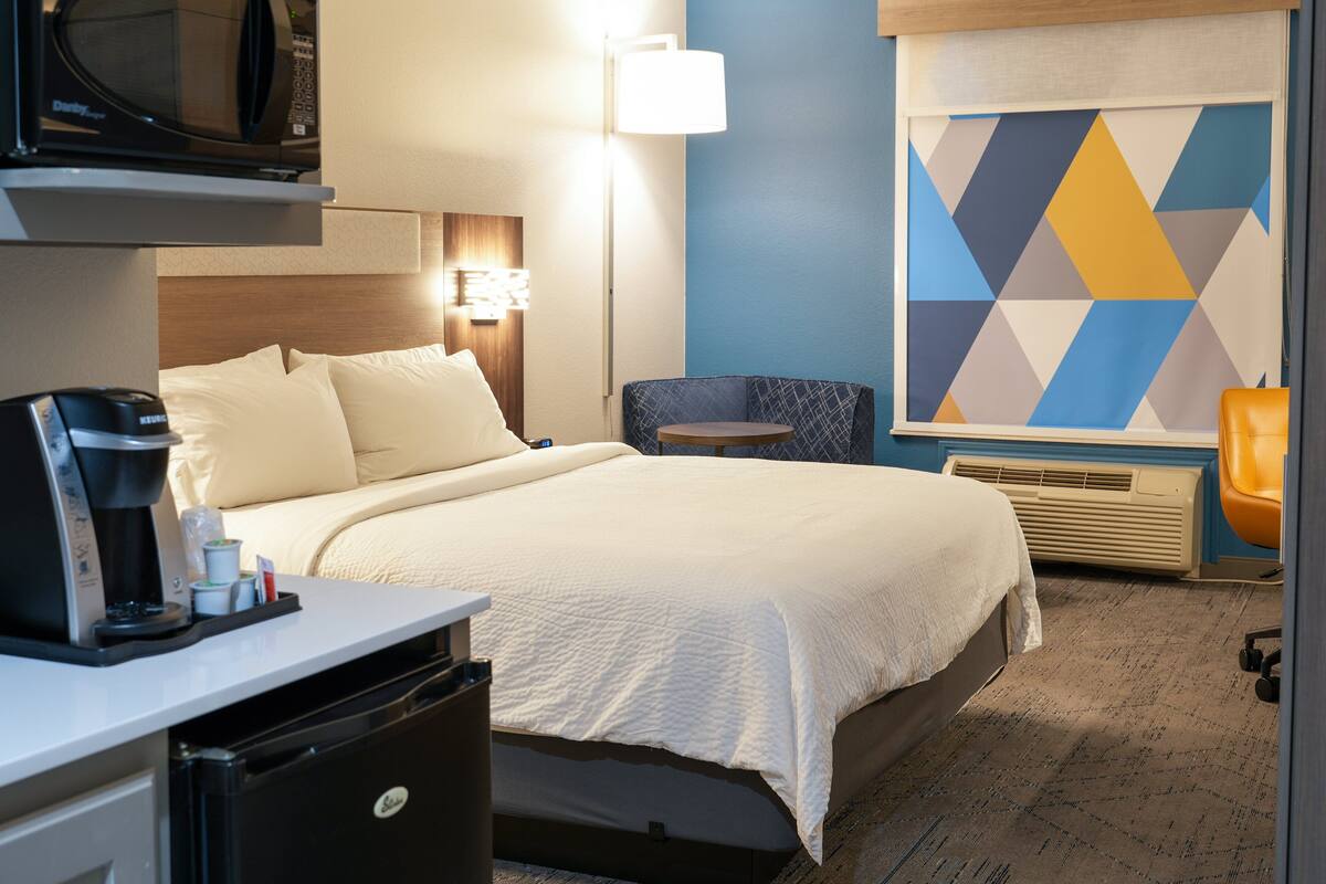 A hotel room with a bed, a desk with a coffee maker, a microwave, a lamp, a chair, and a geometric-patterned wall.