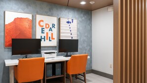 Un espace de travail comprenant deux bureaux, chacun équipé d’un écran d’ordinateur et d’une chaise orange. Des œuvres d’art encadrées ornent le mur, et une imprimante est posée sur les bureaux.