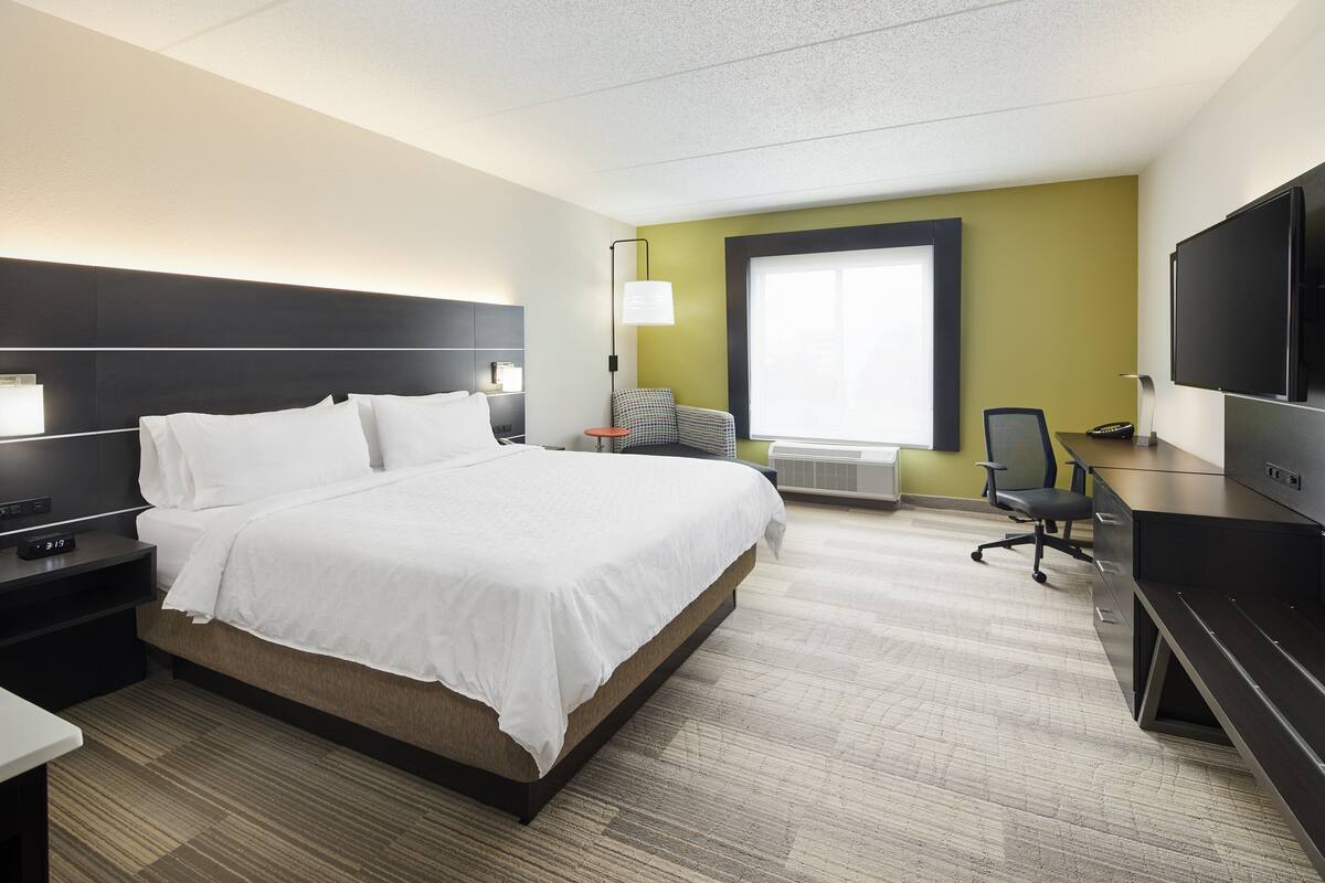 A hotel room with a large bed, a desk with a computer, a chair, a TV, and a window with blinds.