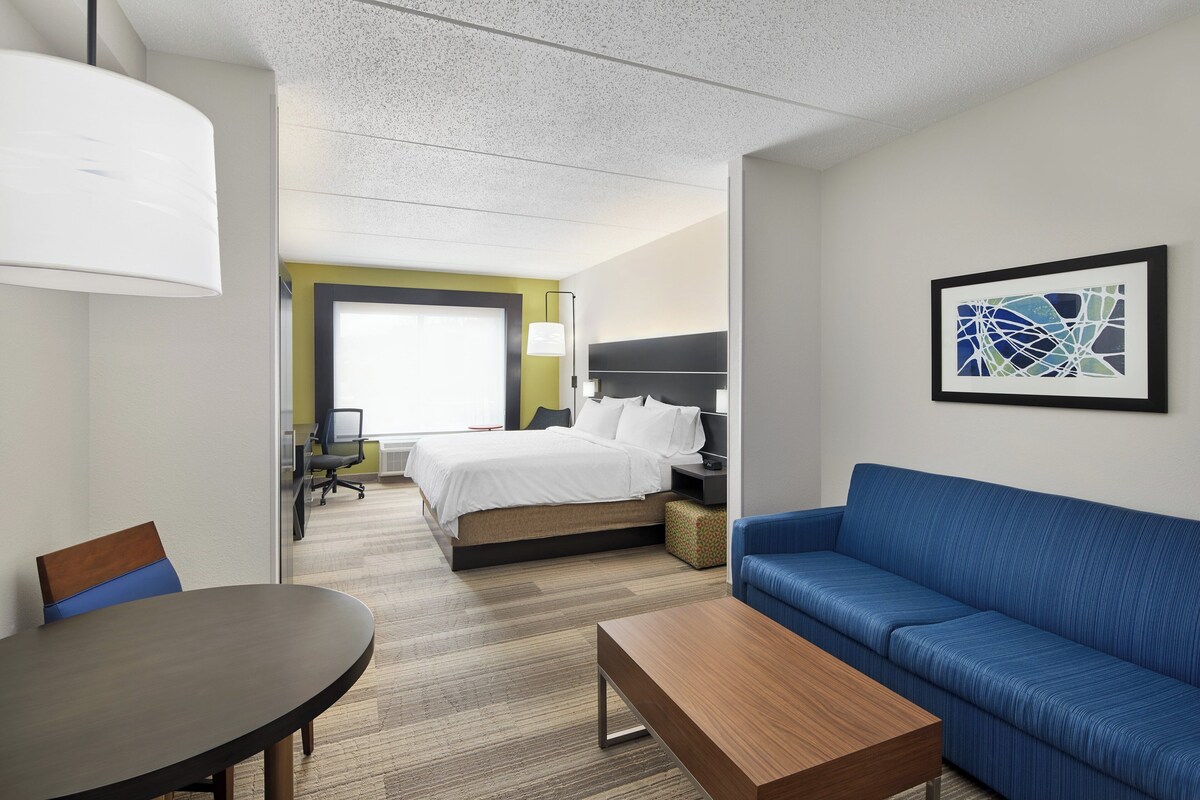 Habitación de hotel con una cama, un sofá azul, una mesa de madera, una lámpara y un cuadro abstracto enmarcado en la pared.