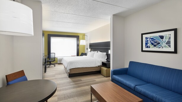 Habitación de hotel con una cama, un sofá azul, una mesa de centro de madera, un escritorio con una silla y un cuadro abstracto enmarcado en la pared.