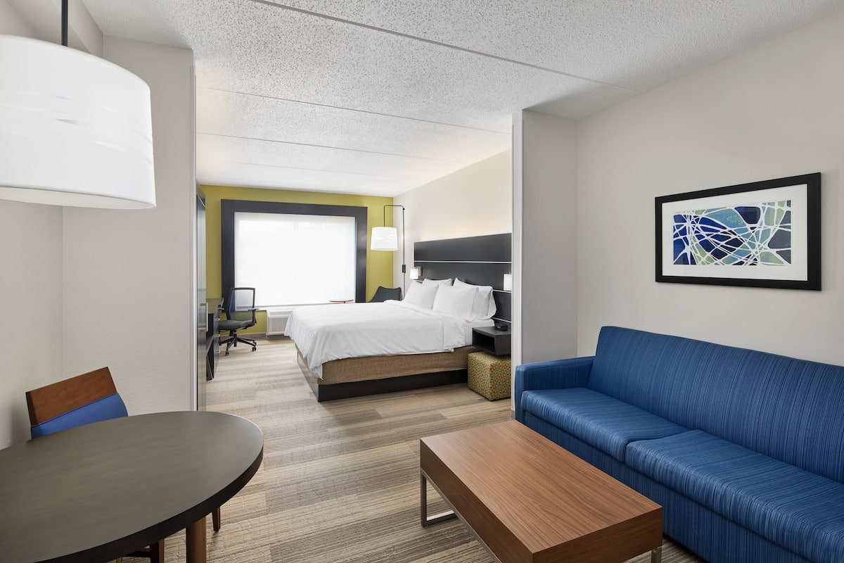 A hotel room with a bed, a blue sofa, a wooden coffee table, a desk with a chair, and a framed abstract painting on the wall.