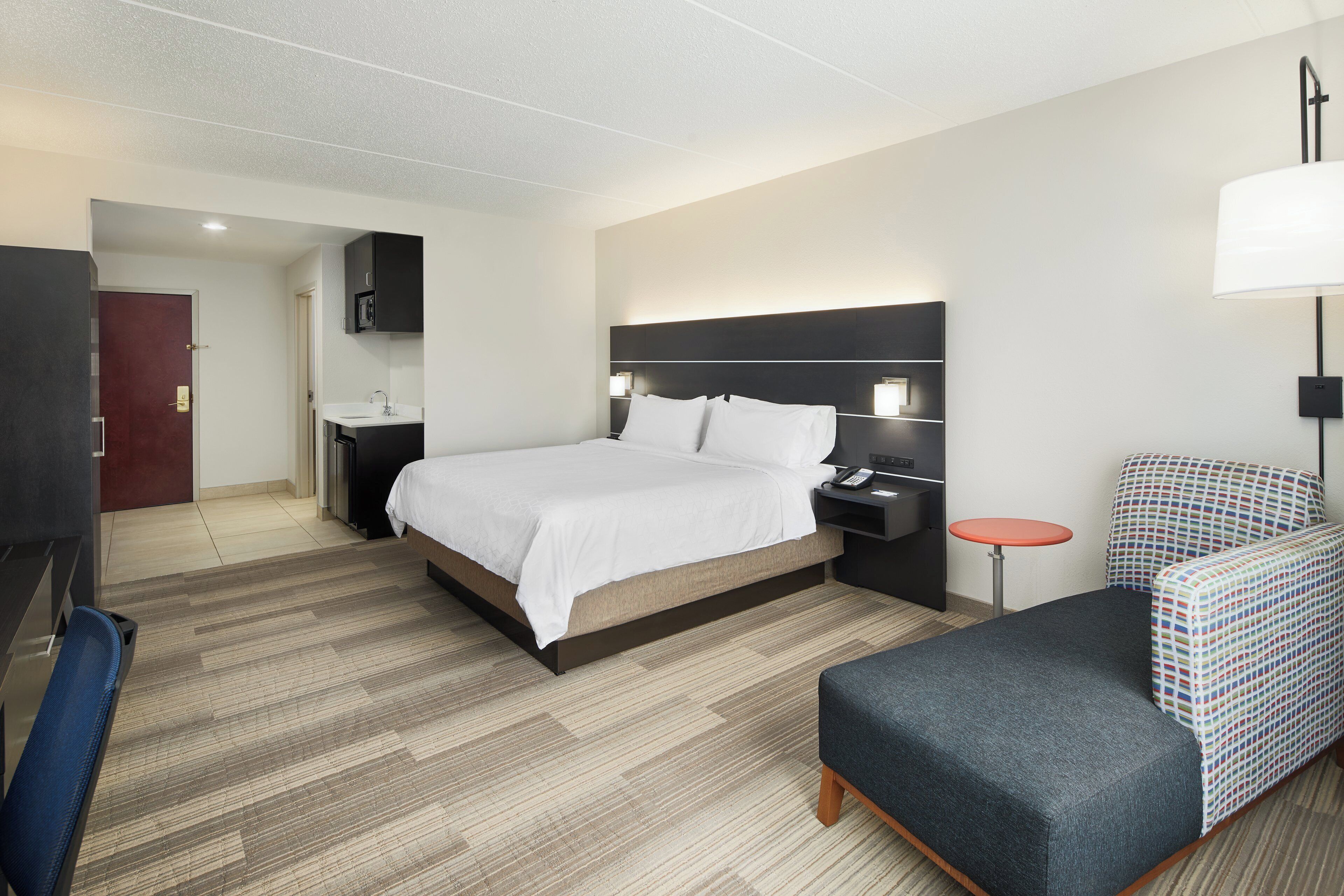 A hotel room with a bed, bedside table, lamp, chair, and a desk with a chair.