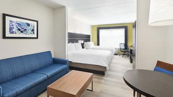 A hotel room with a blue sofa, a wooden table, a bed with white bedding, a desk with a computer, and a large window.