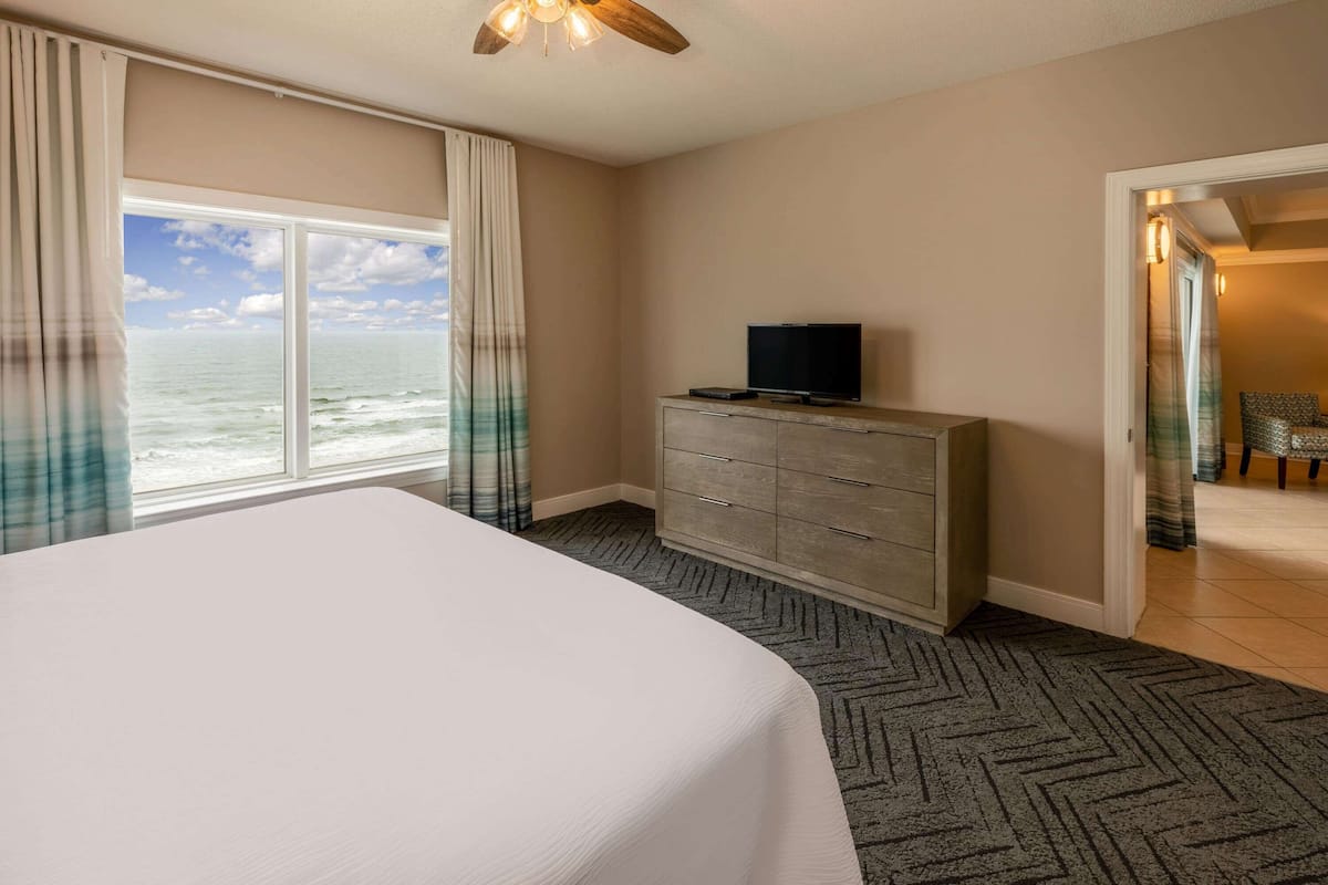 Habitación de hotel con una cama grande, un televisor sobre un tocador de madera y vista al océano a través de un ventanal.