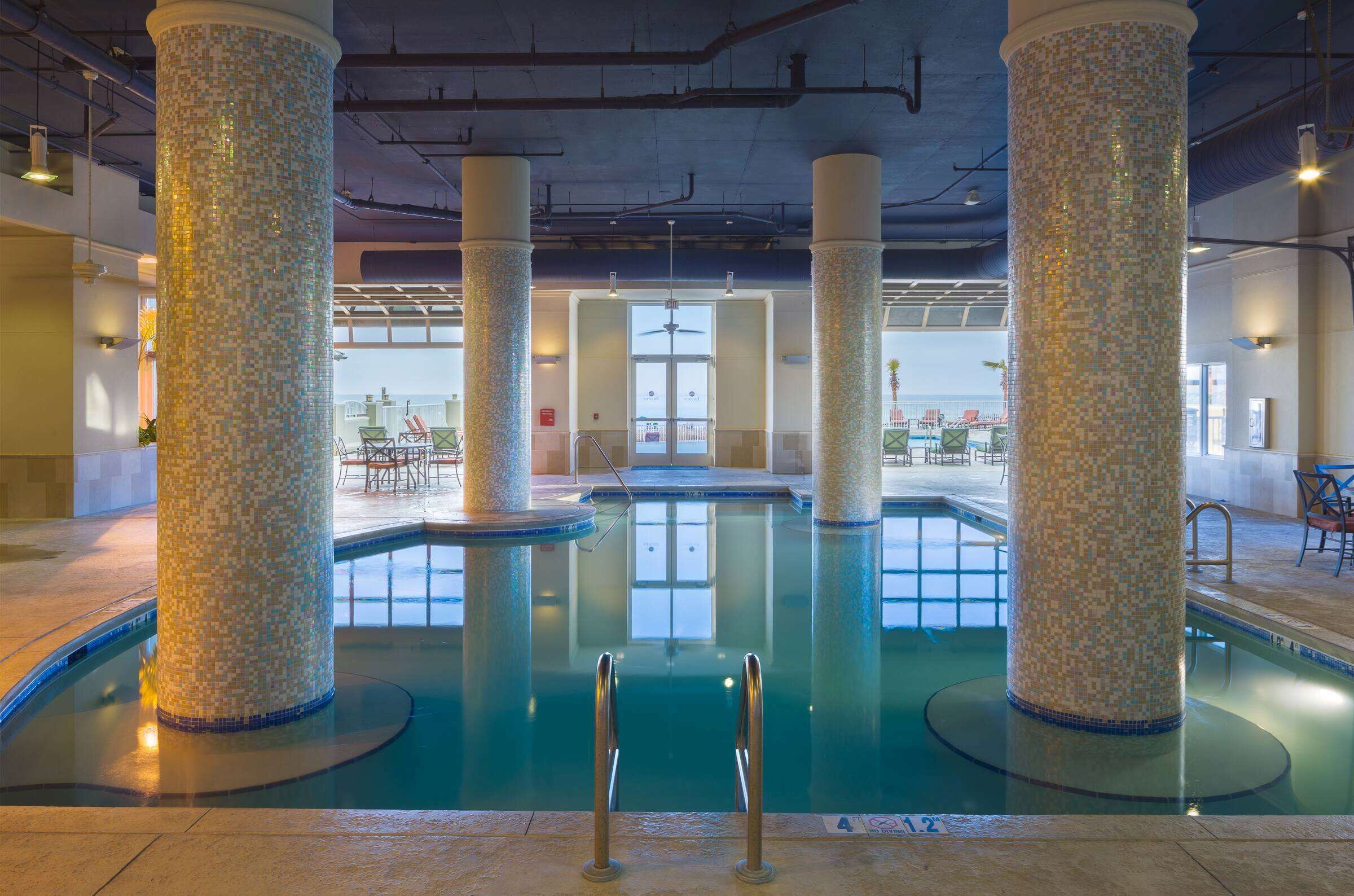 An indoor swimming pool with mosaic columns, clear glass railings, and a view of the outside.