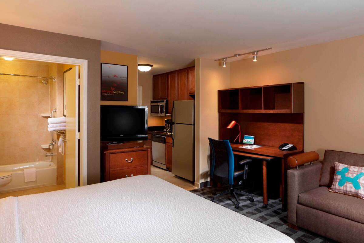 A hotel room with a bed, a desk, a chair, a kitchenette, and a bathroom.