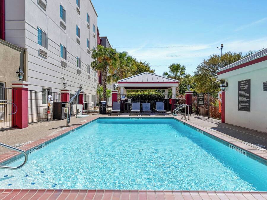 Piscina com um prédio vermelho e branco ao fundo, área coberta com cadeiras e área de piscina cercada.