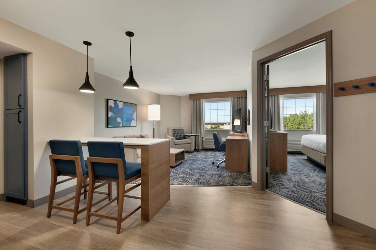 A hotel room with a kitchenette, a dining area with bar stools, a desk, a computer, a bed, and a large window.