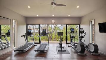 A modern home gym with treadmills, elliptical machines, and a stationary bike, featuring large windows and a ceiling fan.