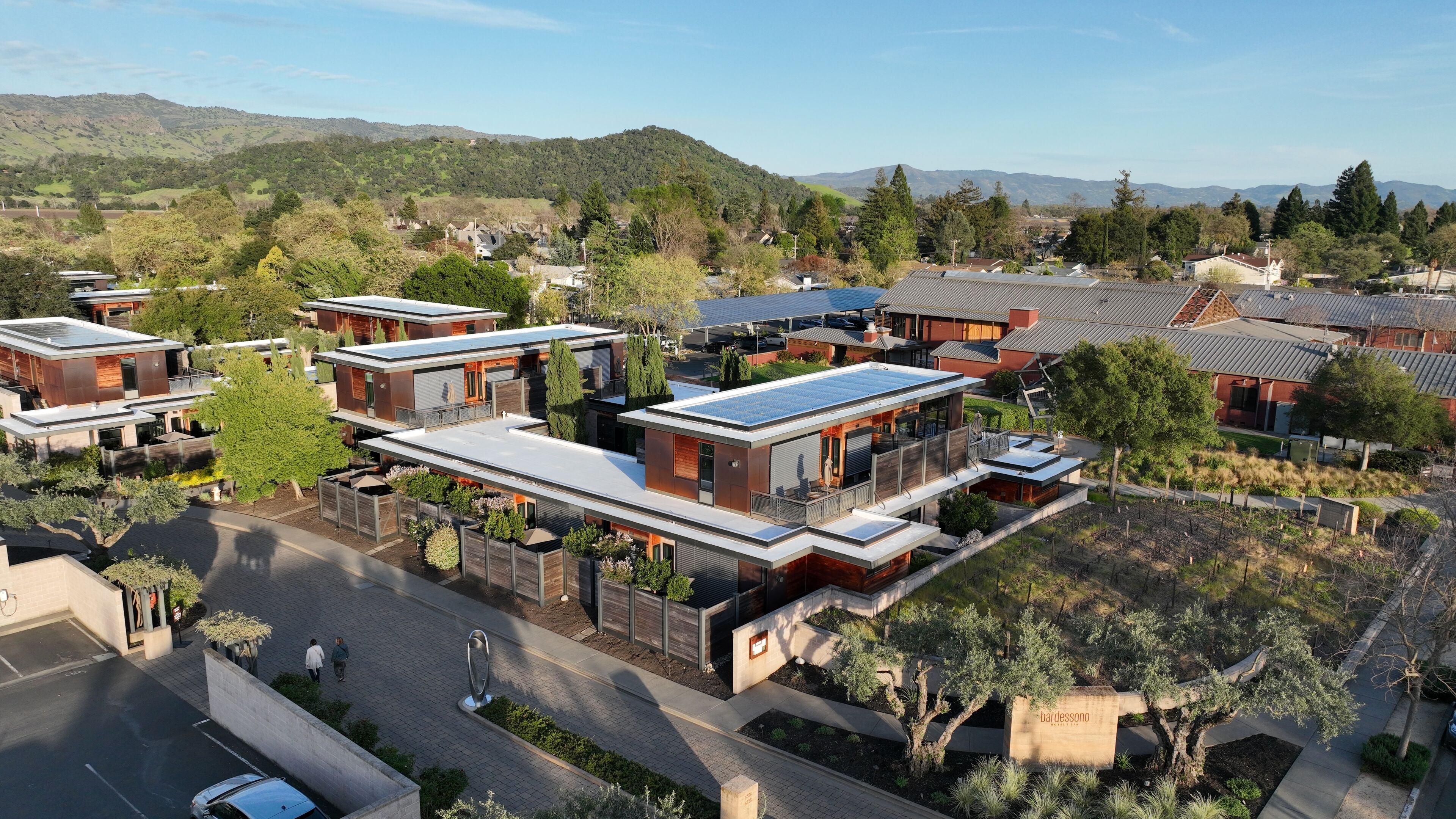 Zona residencial con casas modernas de techos planos y paneles solares, rodeada de vegetación y colinas al fondo.