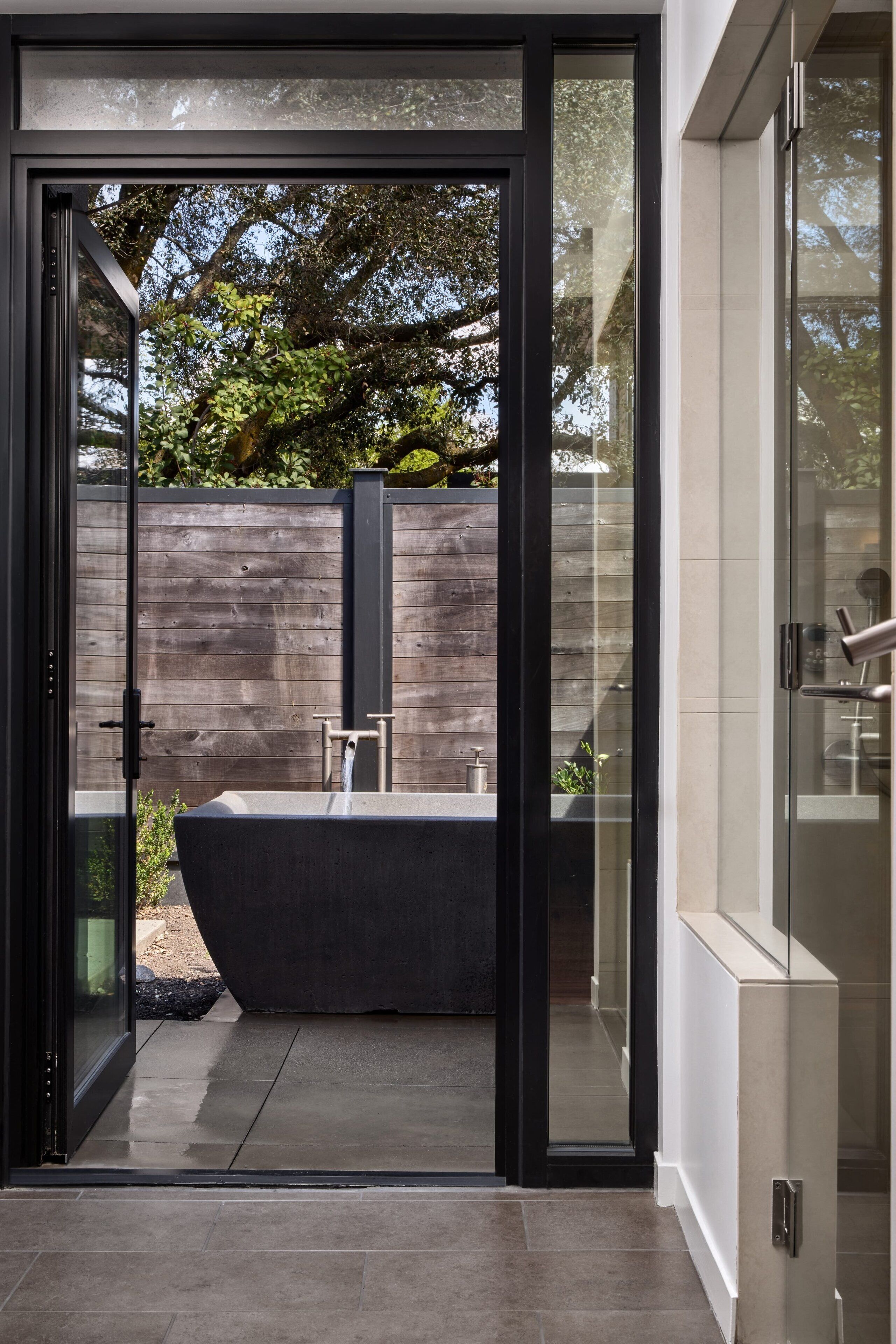 A modern bathroom with a large bathtub, a glass shower enclosure, and a view of a wooden fence and greenery outside.