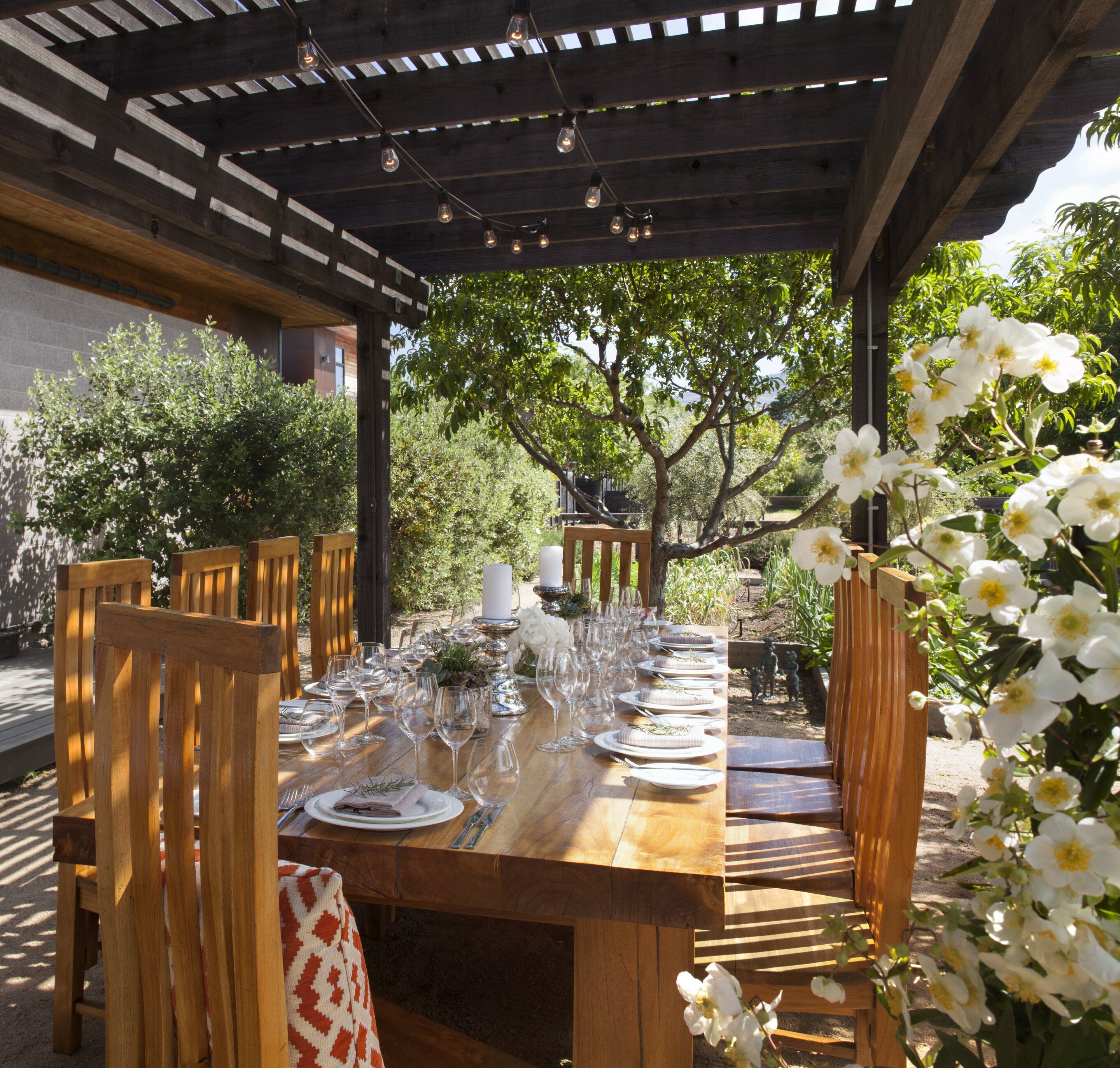 Une pergola en bois installée à l’extérieur, avec une table à manger dressée pour un repas, le tout entouré de verdure et d’arbres.