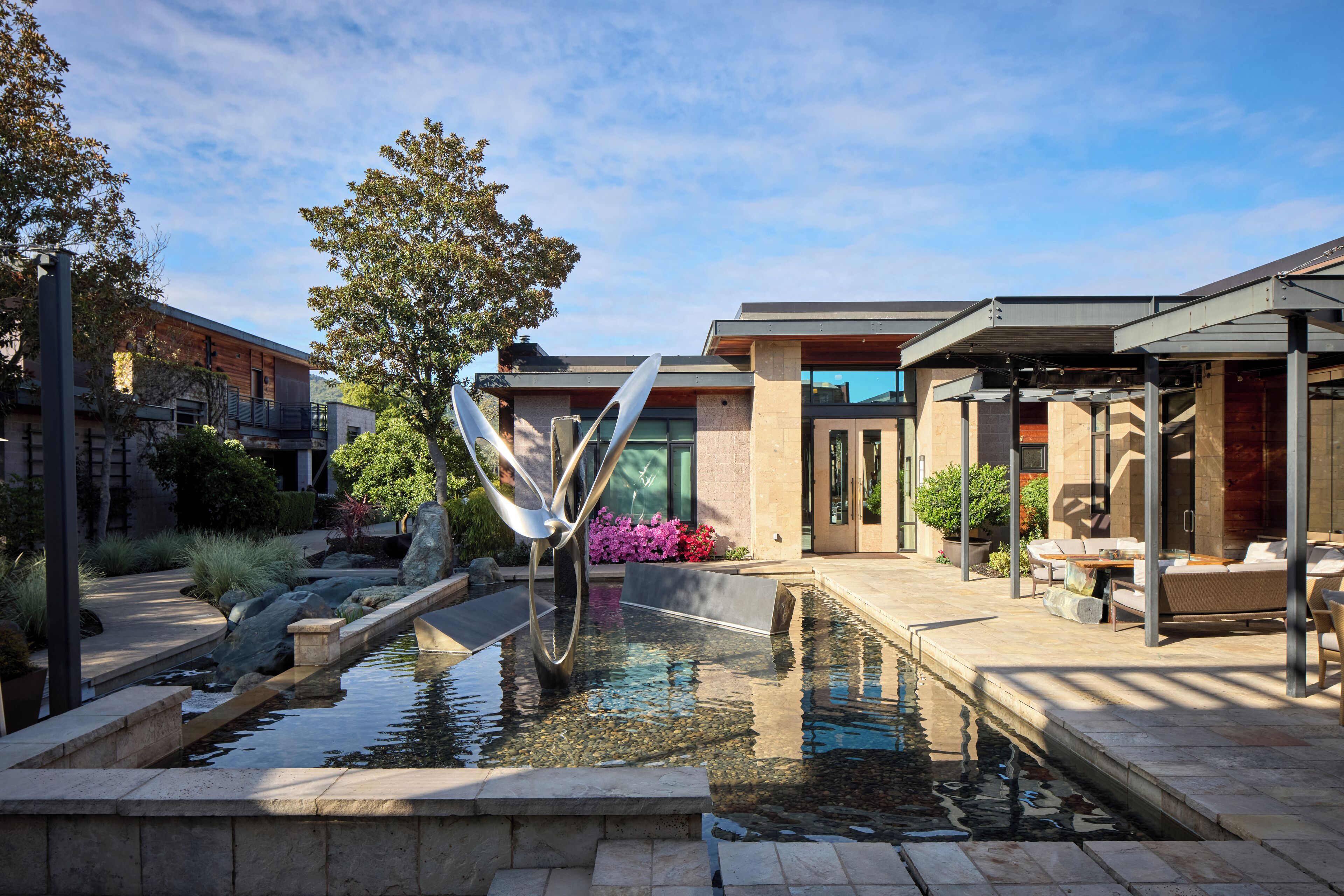 Un área de piscina exterior moderna con una escultura de metal, asientos y un edificio con grandes ventanales.
