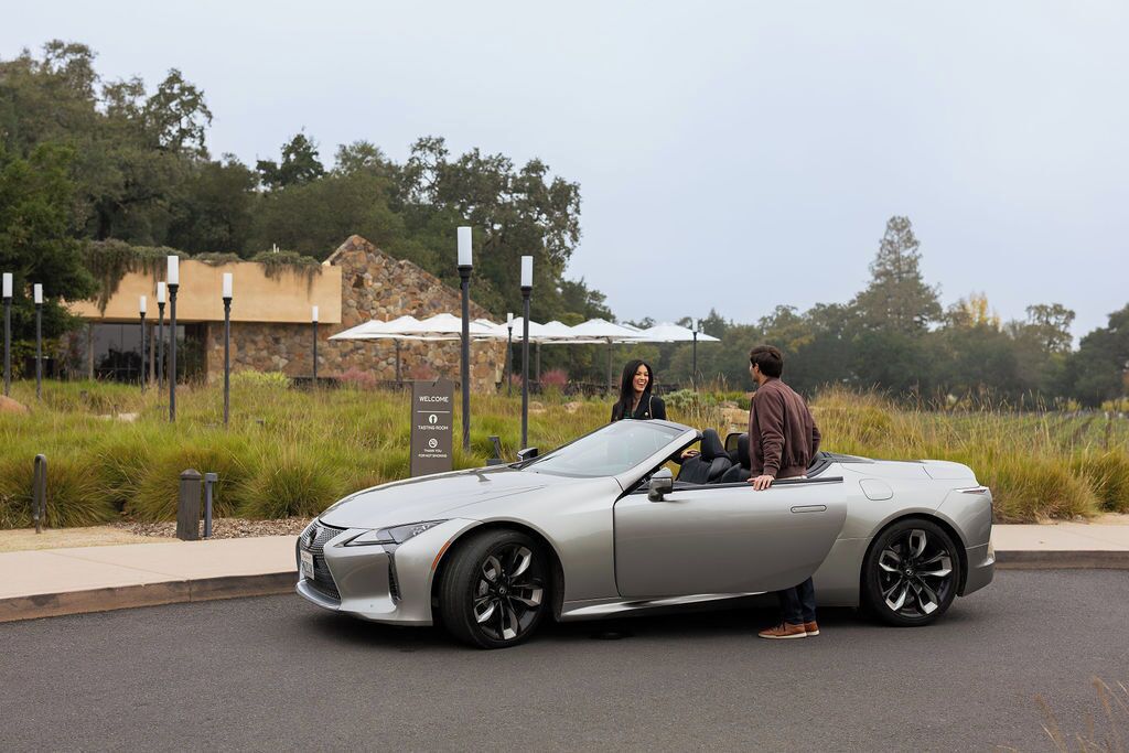 A silver convertible car parked on a road with a sign that reads "Welcome" and "Tasting Room" in the background.