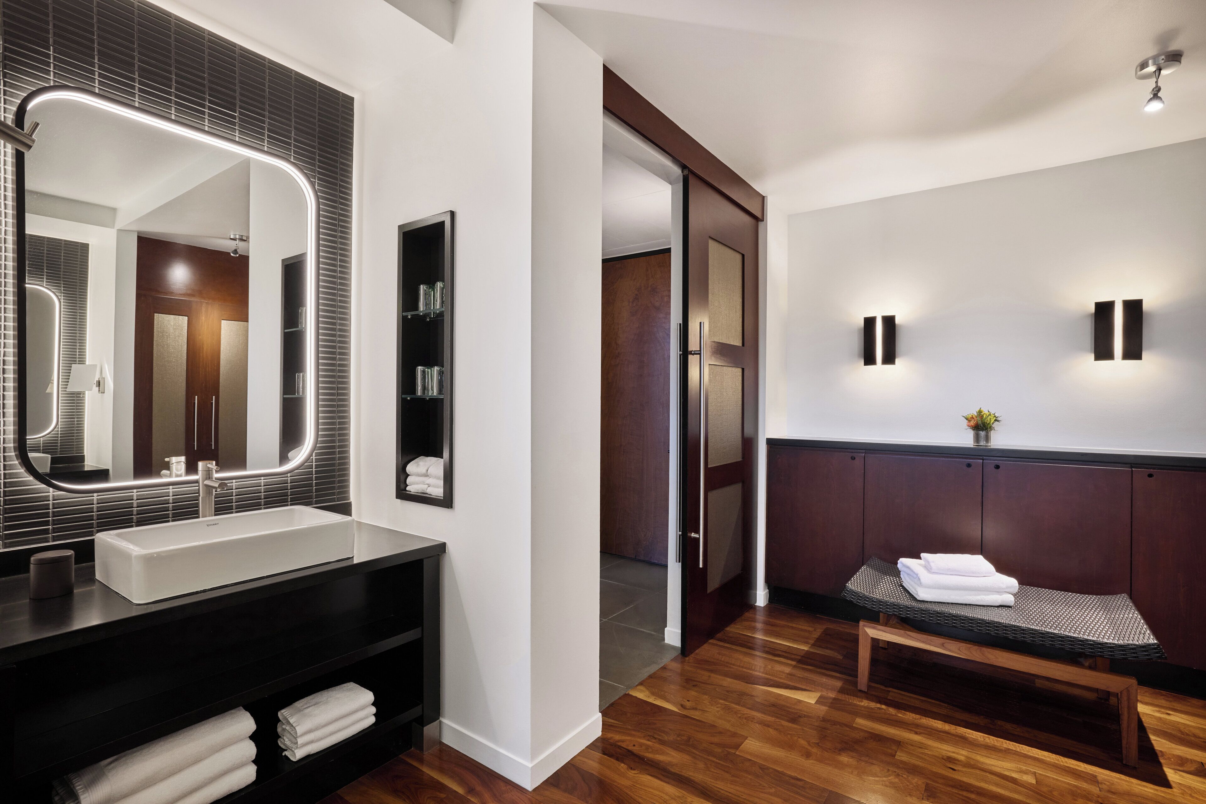 A modern bathroom with a large mirror, a sink, and a bench with towels.