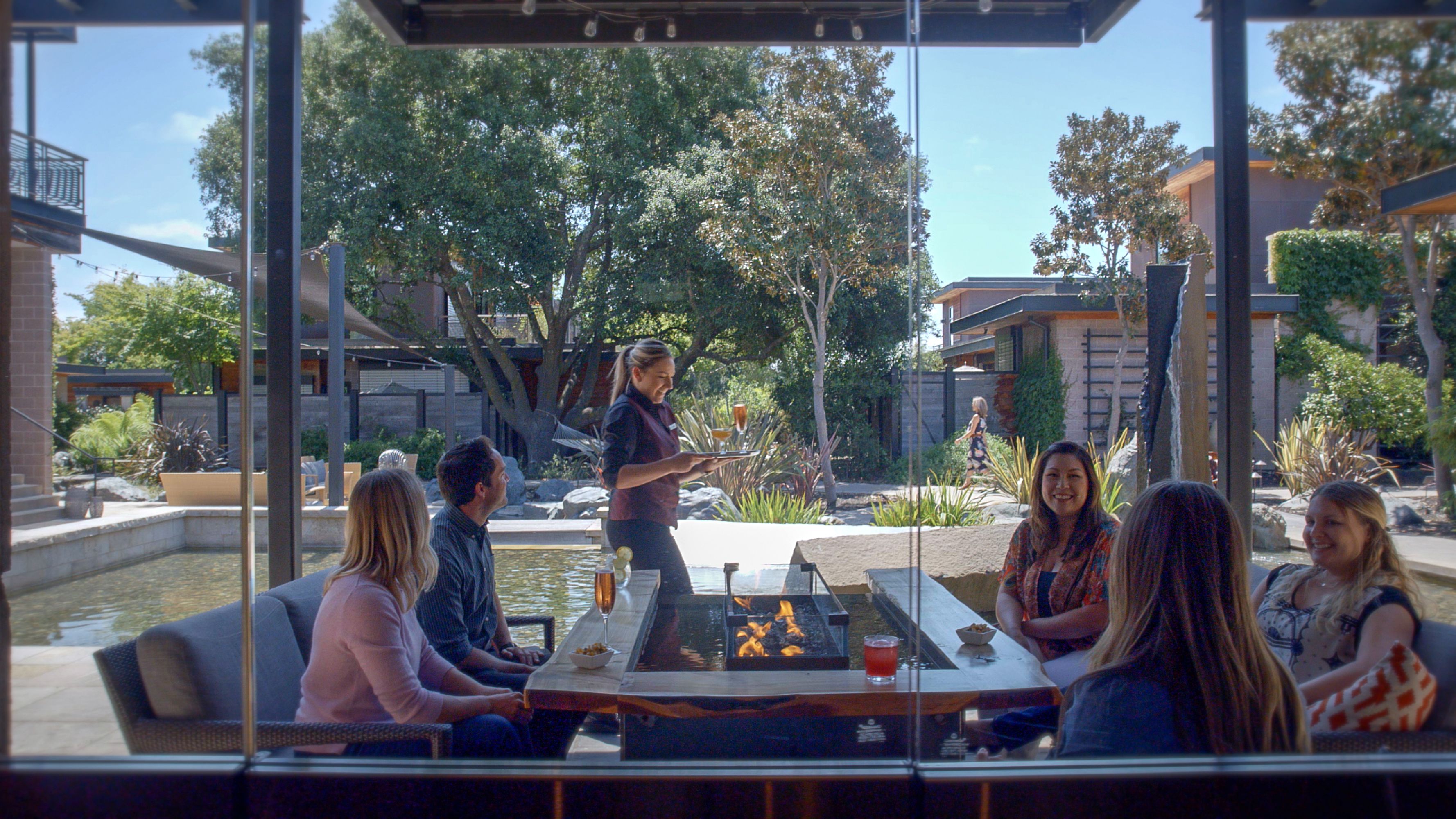 A group of people seated around an outdoor fire pit with a table in between, overlooking a garden area with trees and a water feature.