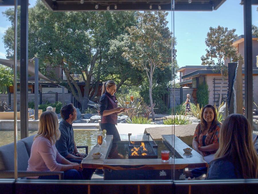 A group of people seated around an outdoor fire pit with a table in between, overlooking a garden area with trees and a water feature.