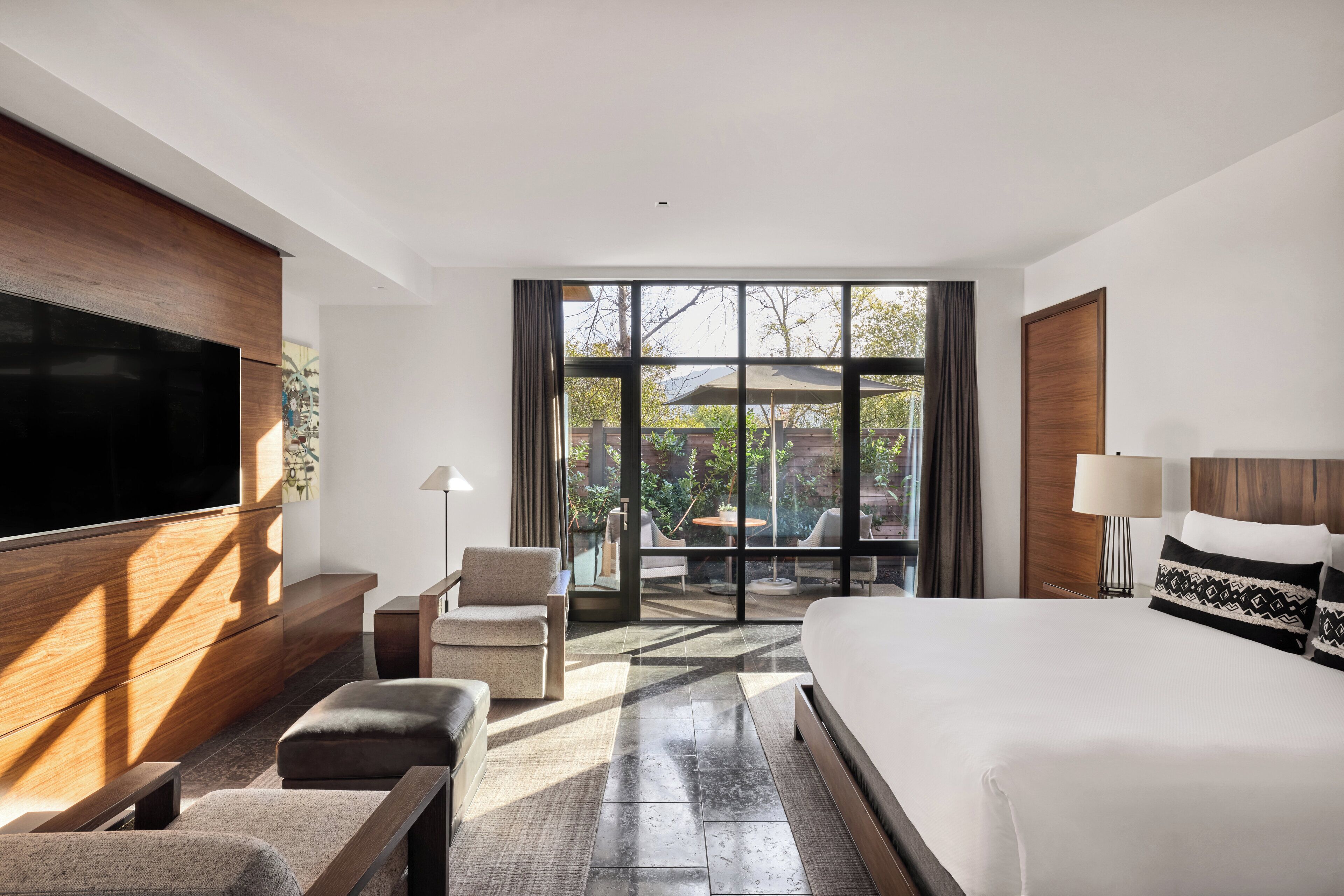 A modern hotel room with a large bed, a flat-screen TV, a sofa, and a balcony with a view of the outdoors.