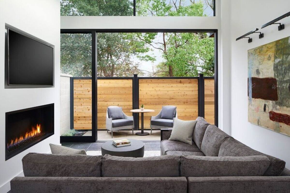 Una sala de estar moderna con chimenea, un sofá grande, dos sillones, una mesa redonda y una terraza de madera visible a través de amplios ventanales.