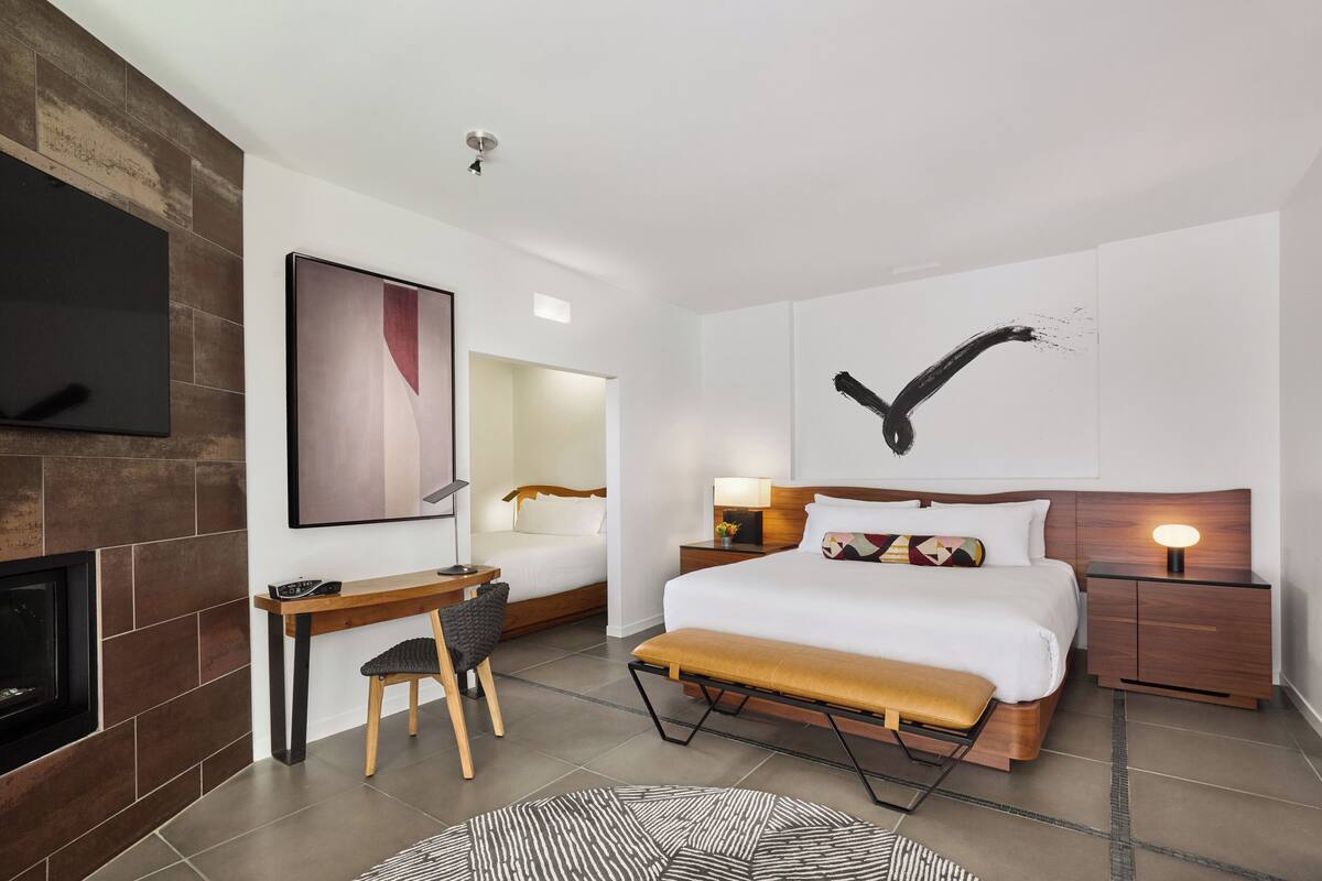 Una habitación de hotel moderna con una cama grande, un escritorio, una silla, un televisor, una chimenea y una obra de arte en la pared.