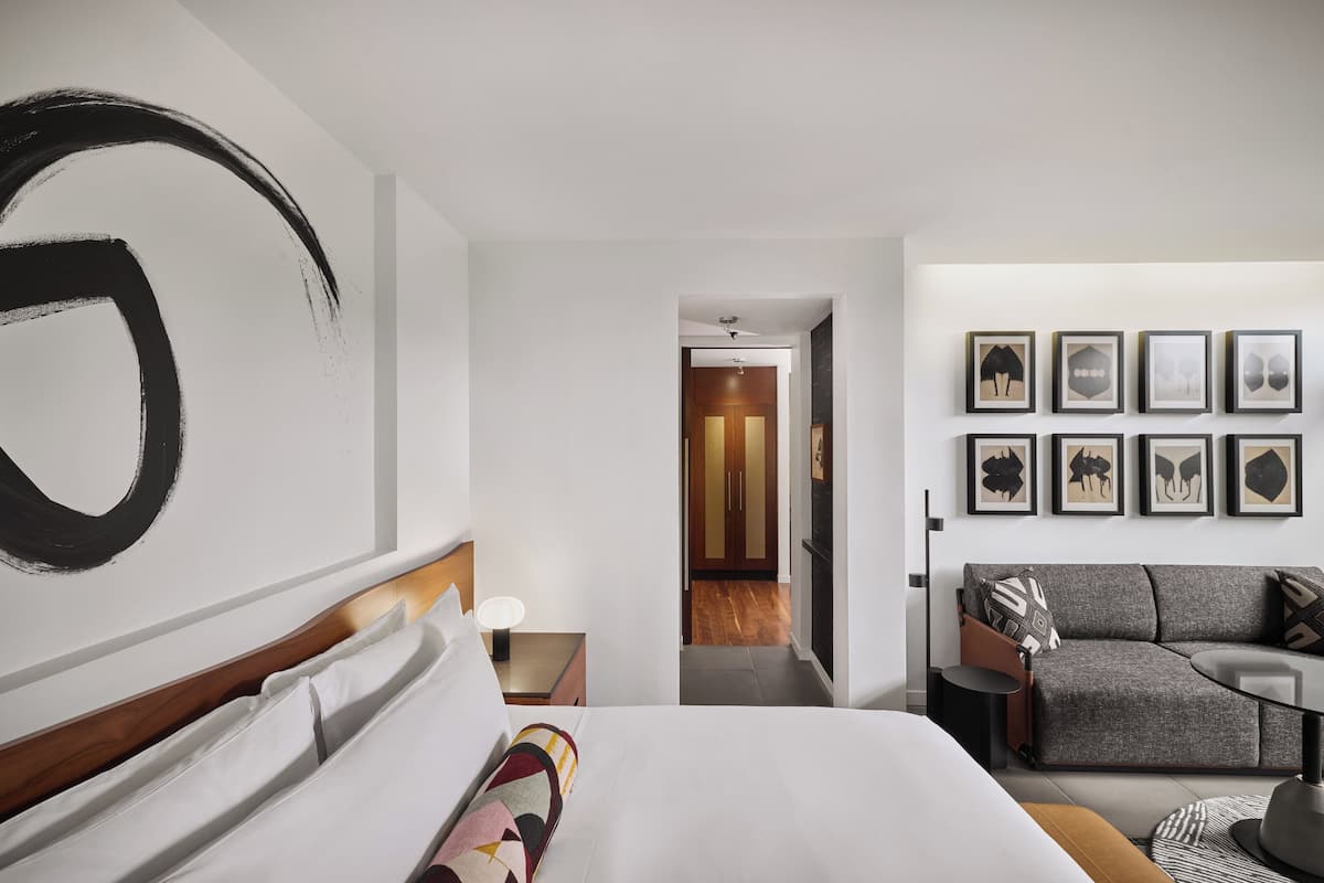 Una habitación de hotel moderna con una cama grande, un sofá gris y un pasillo que lleva a un baño.