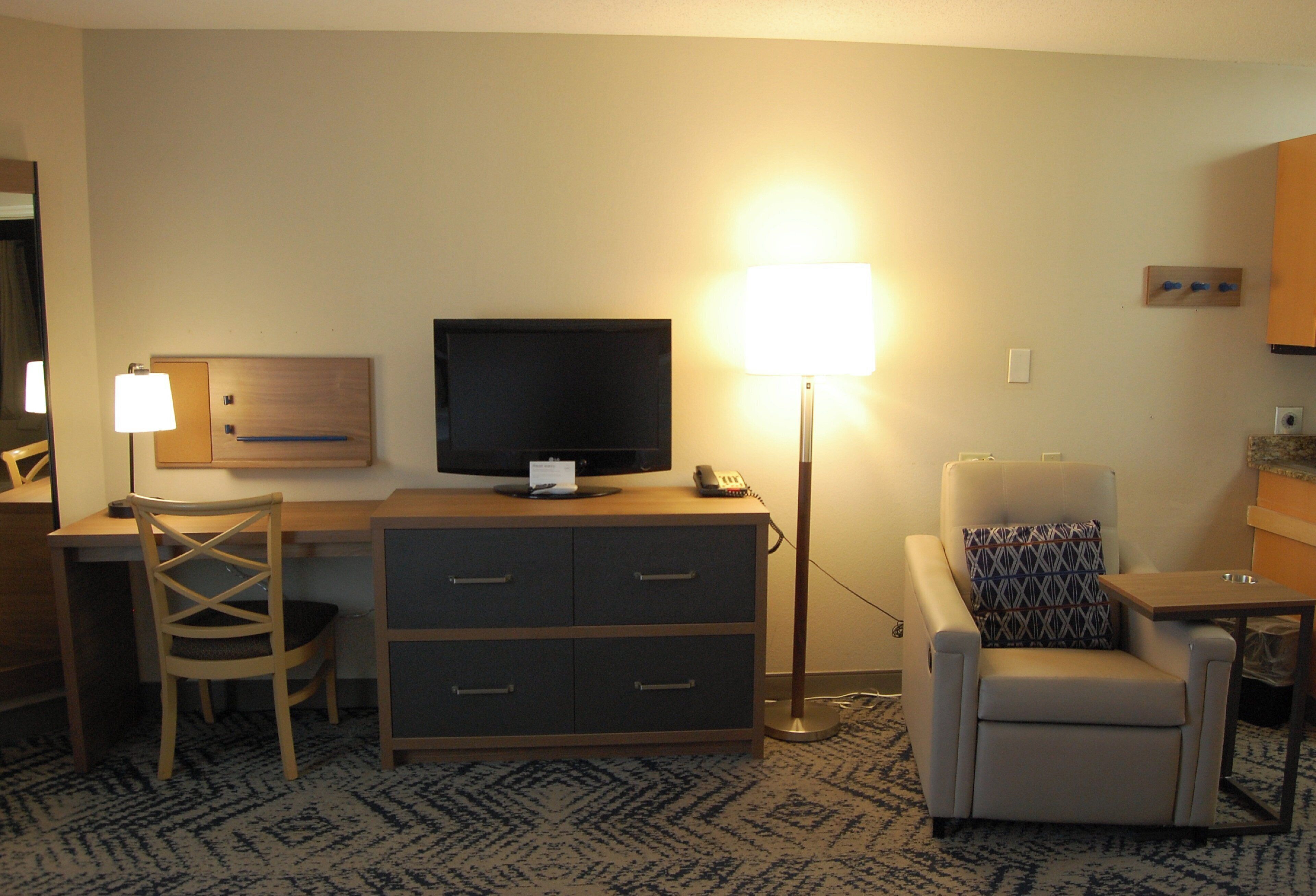 A hotel room with a desk, chair, television, lamp, and a small sofa.