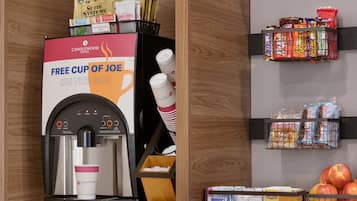 A coffee machine with a promotional sign, a snack shelf, and a fruit display.