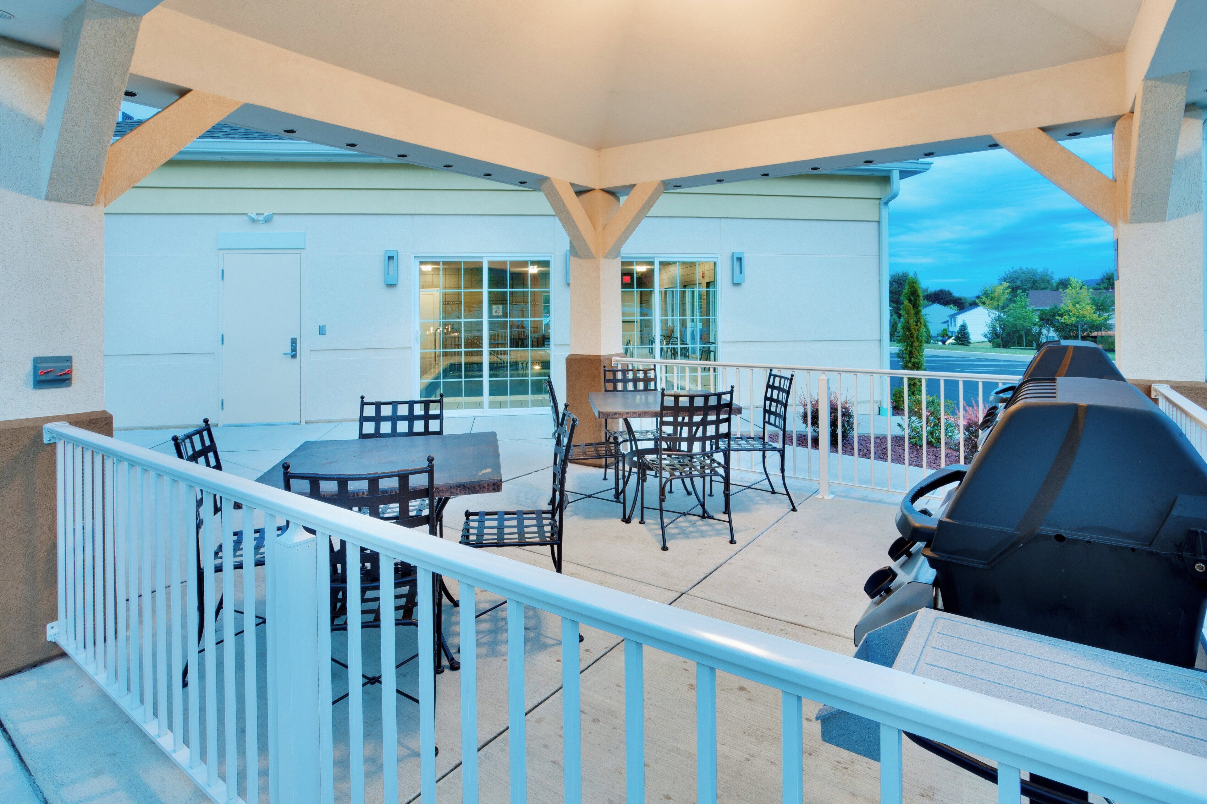 An outdoor patio with a grill, tables, and chairs, overlooking a residential area.