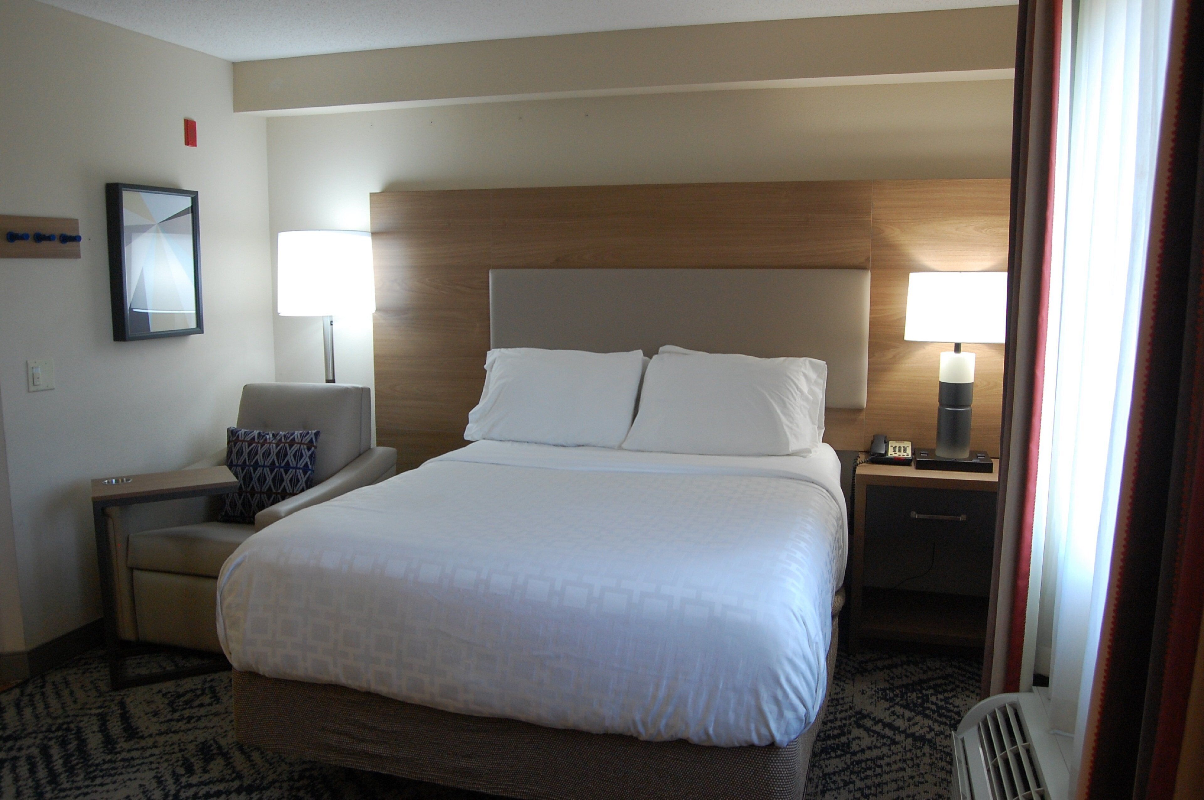 A hotel room with a large bed, a bedside table, a chair, and a nightstand with a lamp.