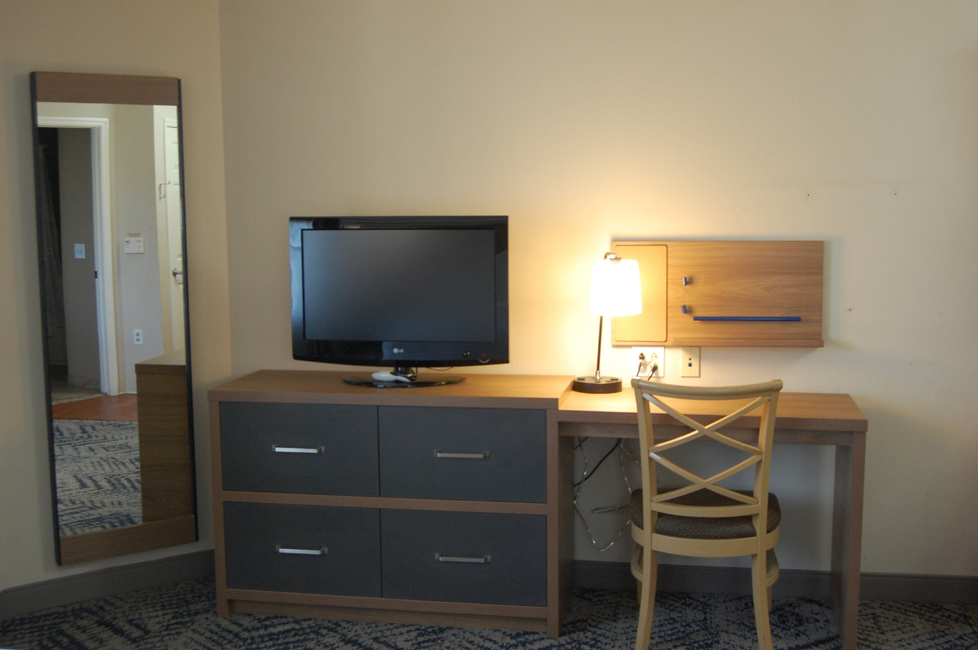 A hotel room with a desk, a television, a lamp, a chair, and a mirror.