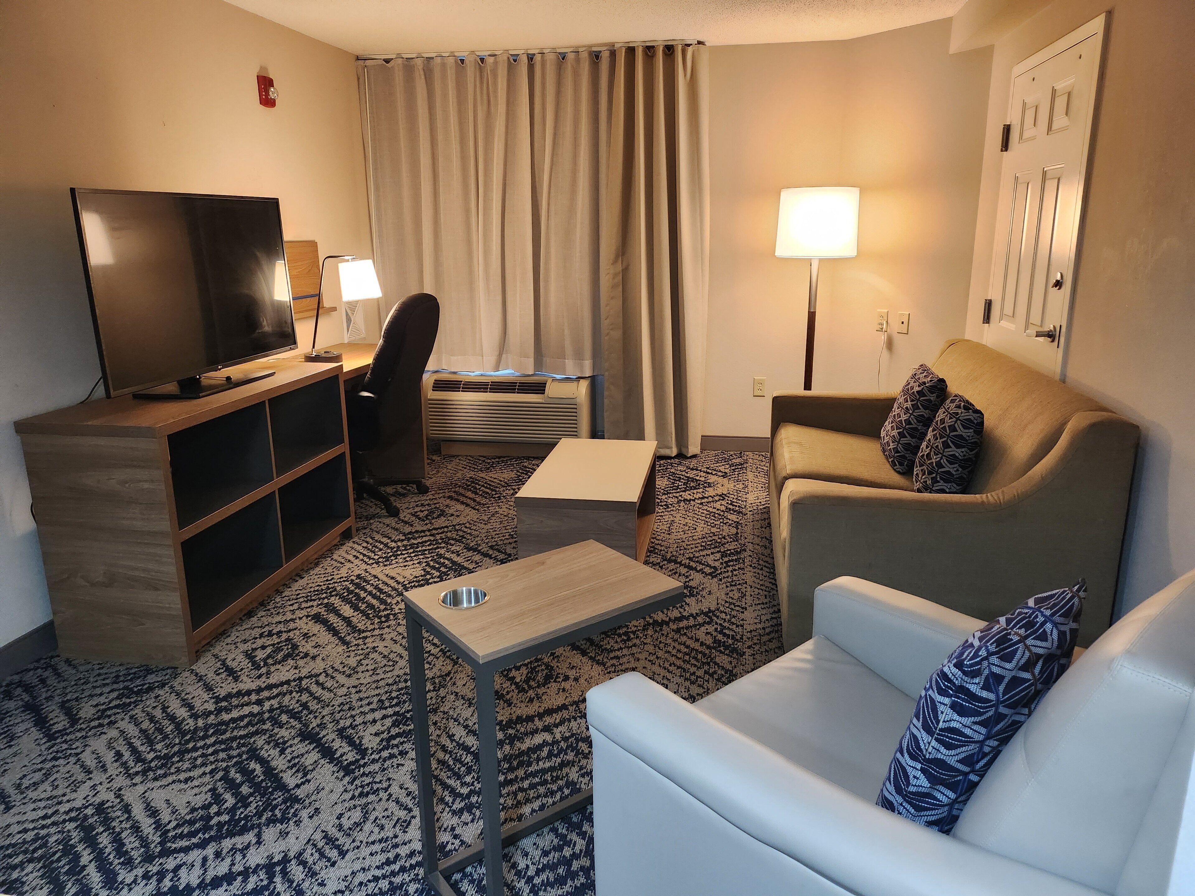 Habitación de hotel con televisión de pantalla plana, escritorio, silla, sofá, lámpara y alfombra estampada.