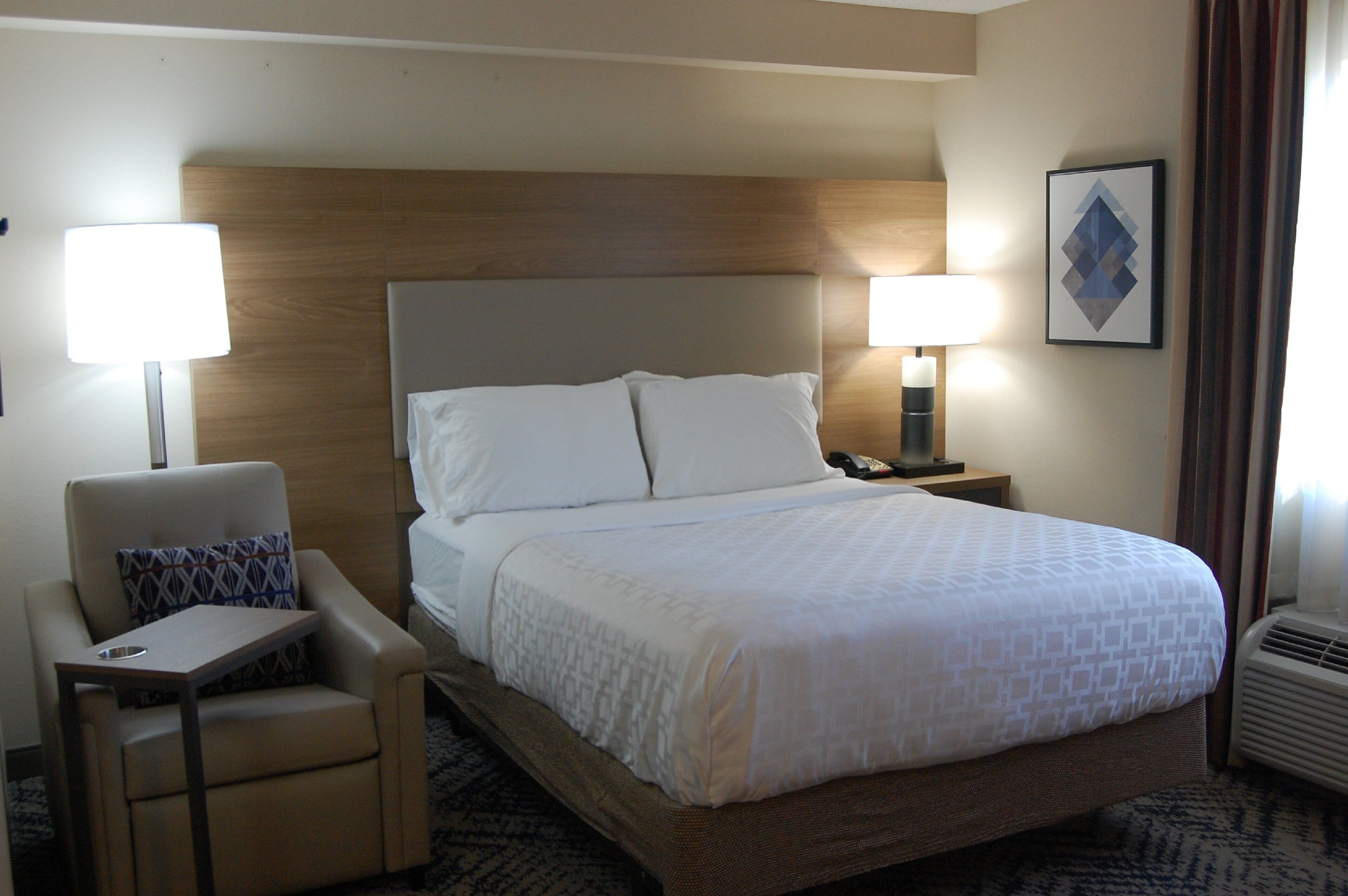 A hotel room with a bed, a chair, two bedside lamps, a phone, and a framed picture on the wall.