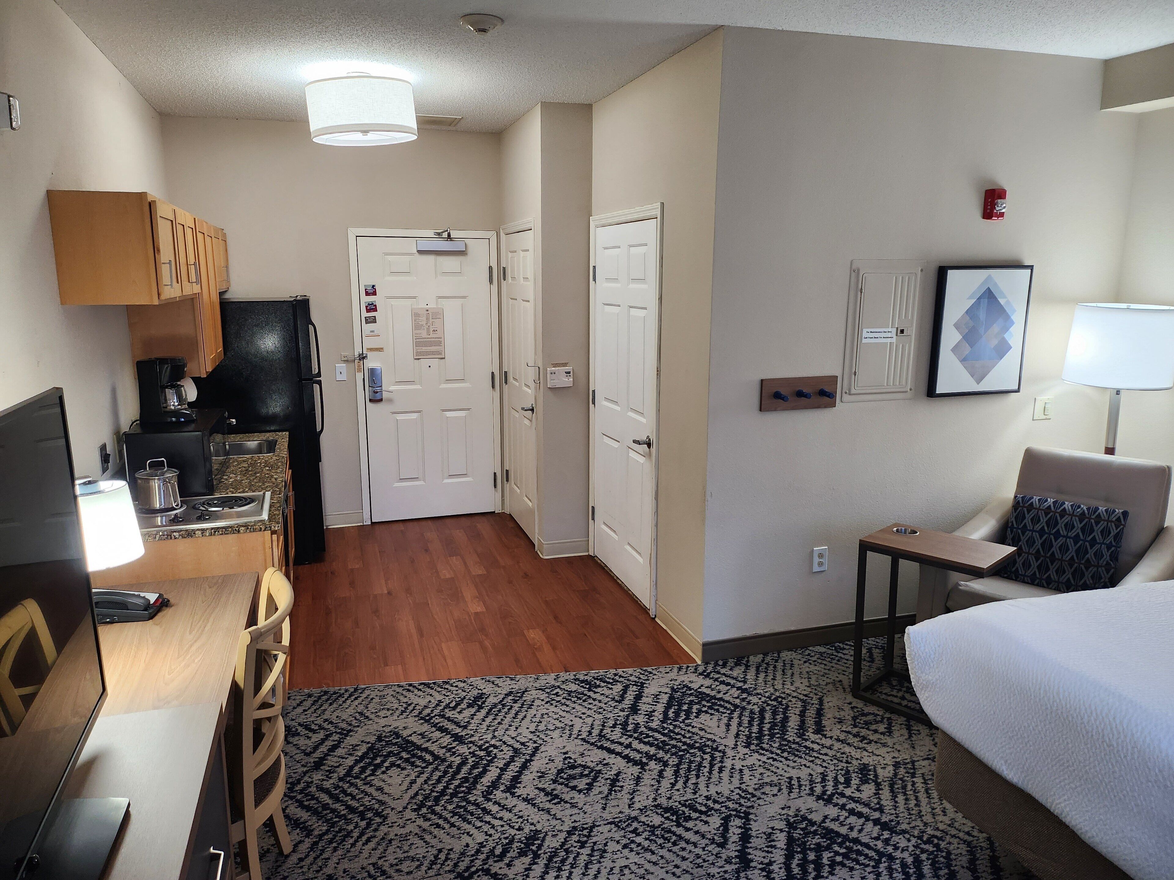 A compact hotel room with a kitchenette, a bed, a desk with a computer, and a seating area with a chair and a table.